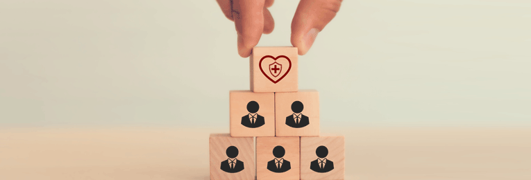 A person arranging cube-shaped blocks with medical logo symbols at the top on a table while cube-shaped human figures are stacked at the bottom.